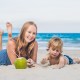 Mom and child laying on stomachs on beach with a coconut in front of them. This articles asks: Does coconut water help with breast milk supply?